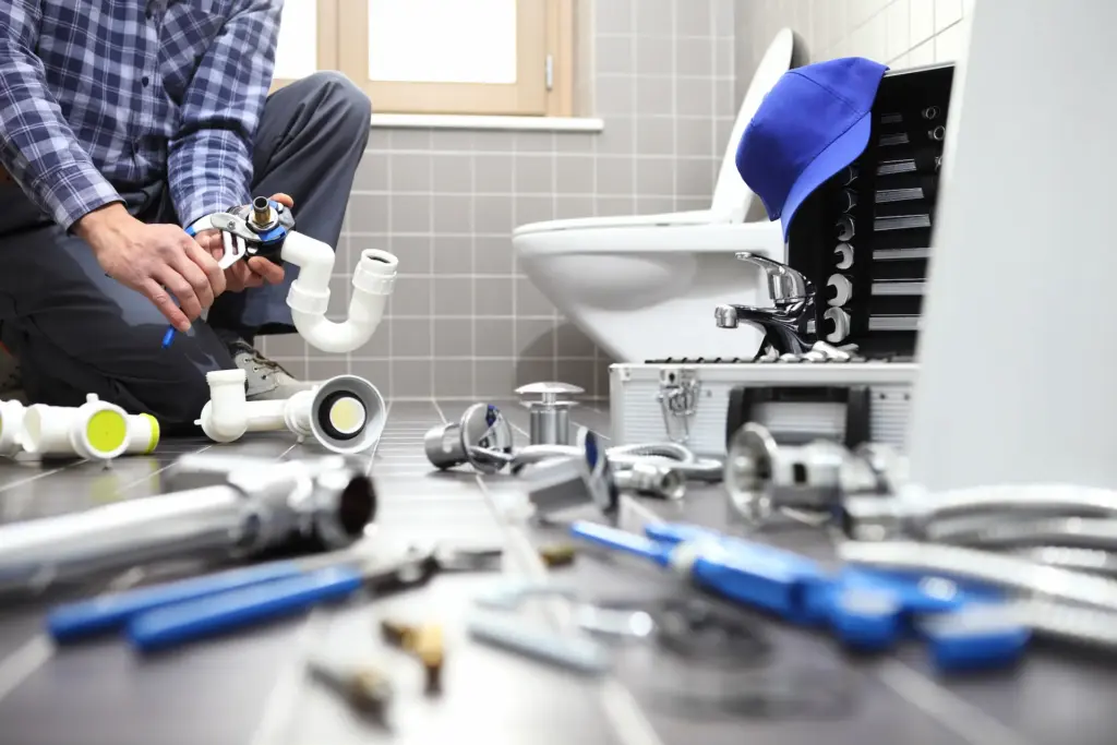 Plumber installing a new toilet in a bathroom with grey tile. His tools are all over the floor and he is piecing together PVC pipes